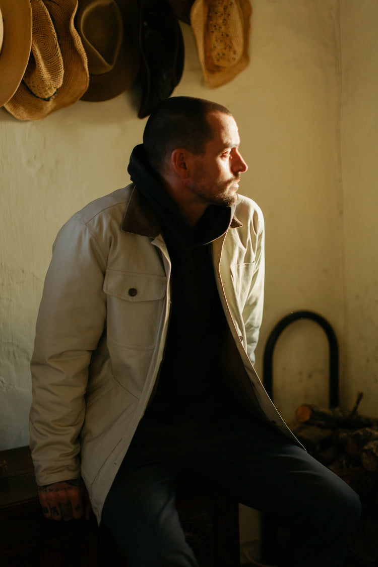 A man sitting in a sunlit cabin wearing the cream insulated Rancher jacket layered over a black Sur hoodie, styled with a western heritage aesthetic.