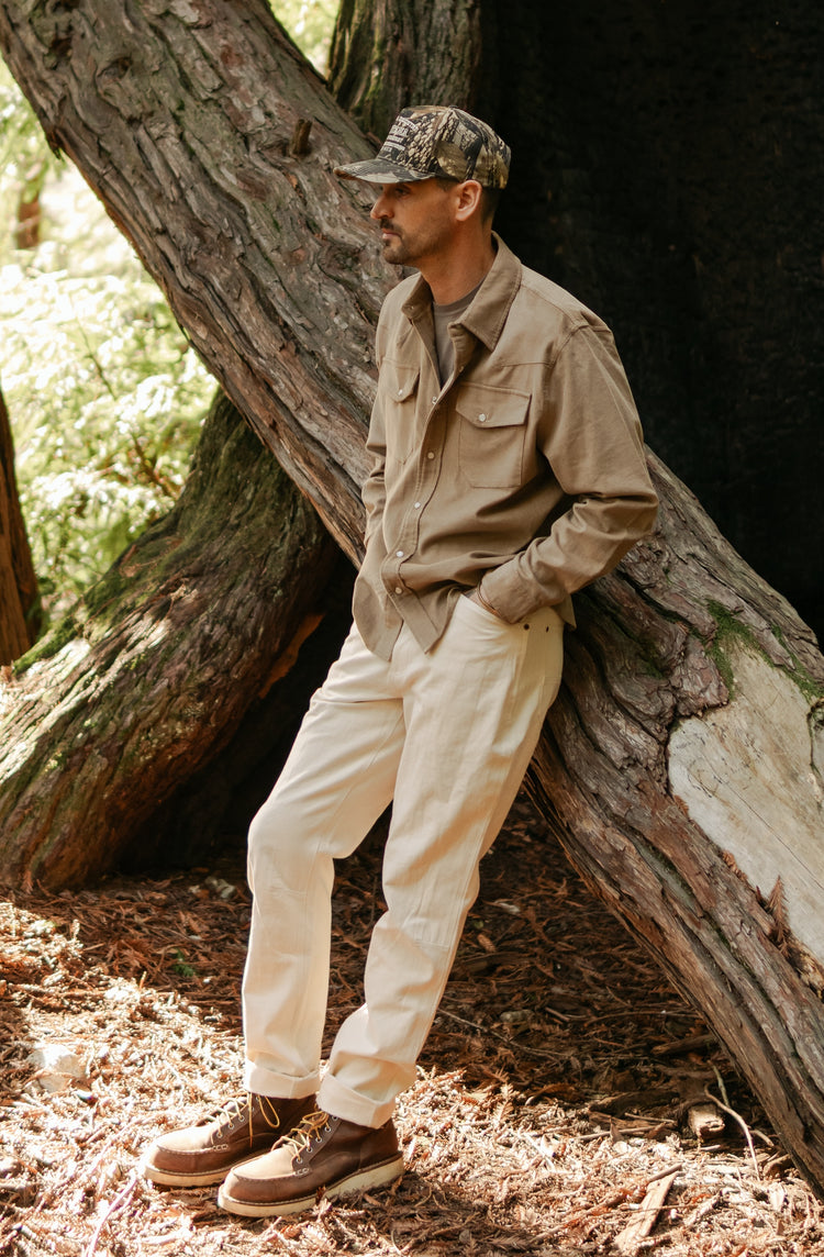 Side profile of the Tradesman Pant in softwashed cream cotton canvas, highlighting the durable triple-needle stitching and straight-leg fit, styled with a brown Western Cord shirt in a forest setting.