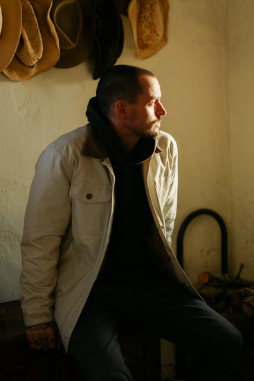 A man sitting in a sunlit cabin wearing the cream insulated Rancher jacket layered over a black Sur hoodie, styled with a western heritage aesthetic.