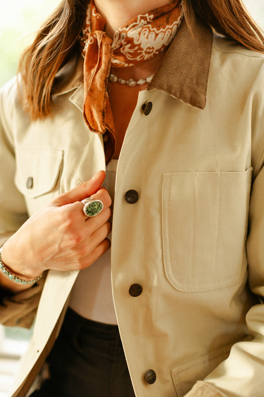 Close-up of a women's cream canvas chore coat featuring a brown corduroy collar, bronze metal buttons, and a reinforced front utility pocket.