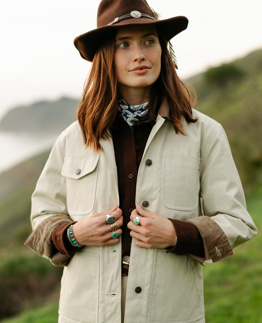 Women’s cream canvas chore jacket with corduroy-lined cuffs and front utility pockets styled with a brown felt hat.