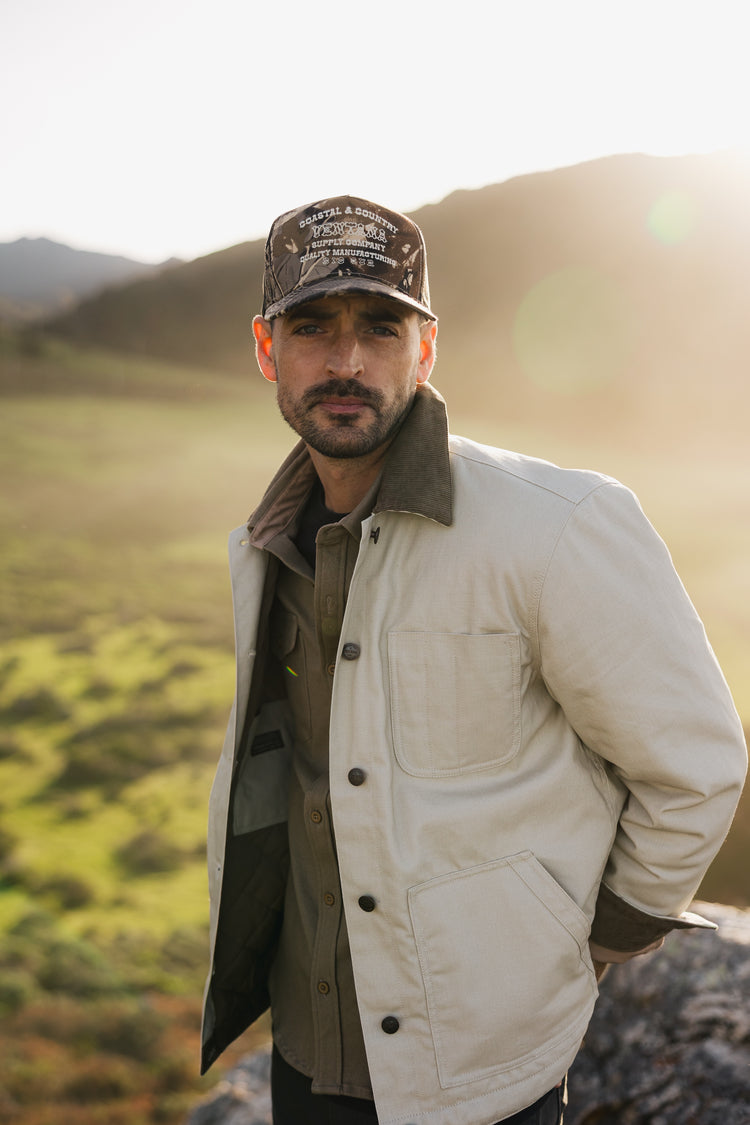 Men’s cream canvas rancher jacket with a unisex fit, featuring a corduroy collar and styled with a camo trucker hat in an outdoor setting.