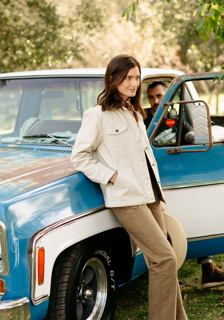 Woman wearing the cream canvas chore jacket and Lily Pant in tan, leaning against a vintage blue truck for a rugged outdoor western look.