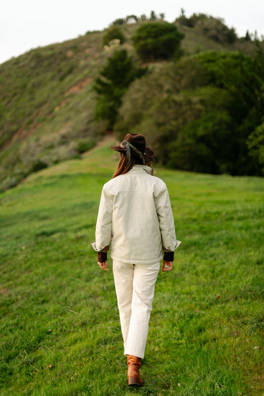 Full-length back view of a woman wearing a cream canvas chore jacket and matching cream Lily Pants for a monochromatic outdoor look.