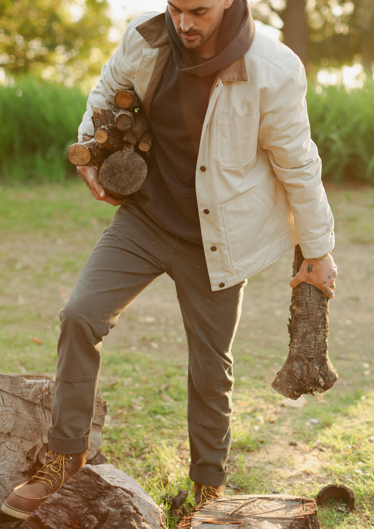 Man carrying firewood wearing a cream insulated Rancher jacket, a faded black Sur hoodie, and charcoal grey Draftsman pants in salt washed coal.