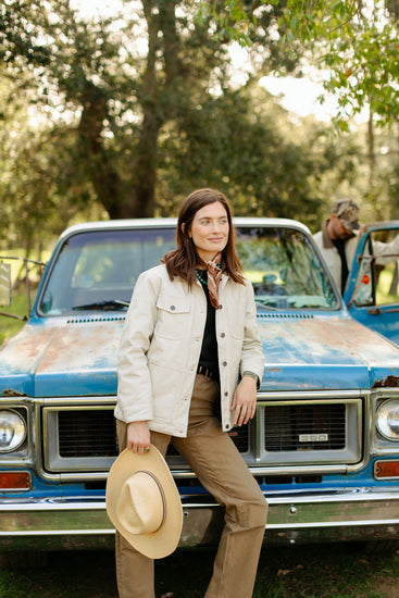 Woman wearing a cream canvas utility jacket leaning against a vintage blue truck, styled with brown work pants and a straw cowboy hat.