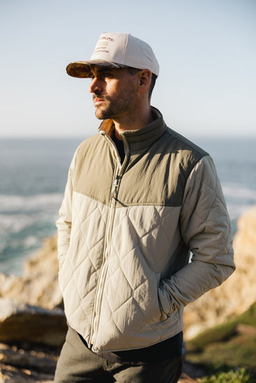 Men’s two-tone Cruzer jacket in sage and cream paired with a corduroy and camo trucker hat, featuring diamond quilting and a stand-up collar.