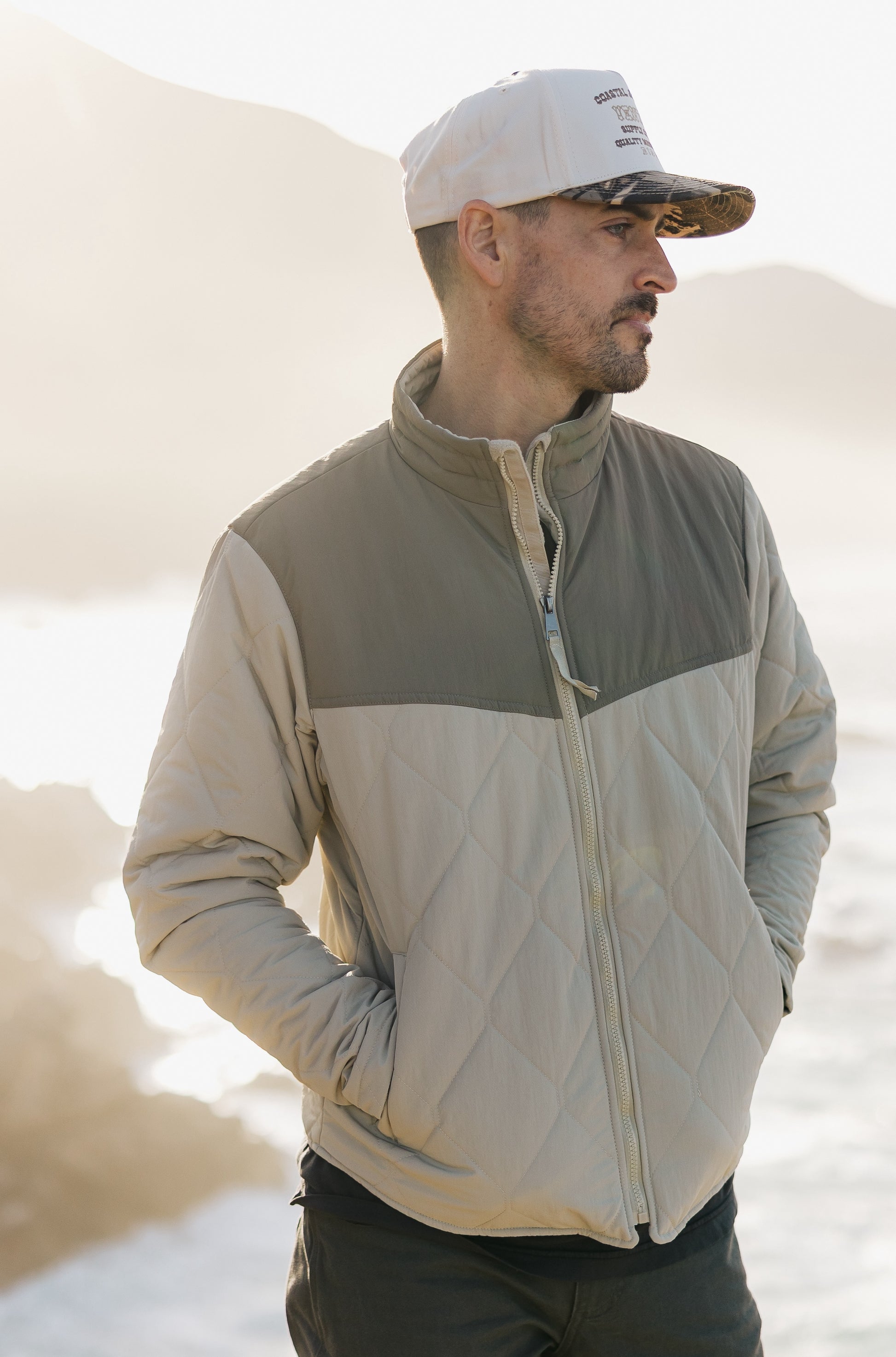 Profile view of the men's Cruzer jacket in sage and cream, paired with a two-tone camo 5-panel hat featuring a cream corduroy crown and camouflage brim.