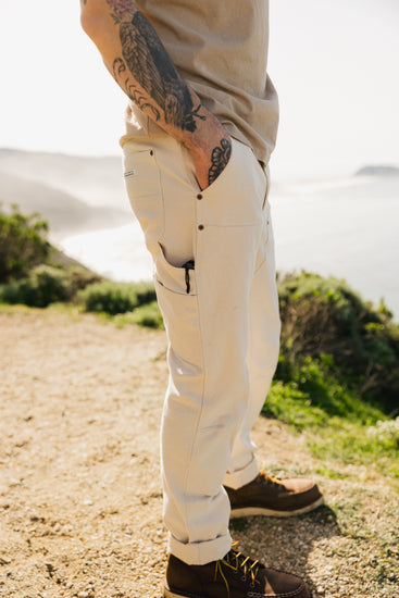 Side view of the VNTNA Draftsman Pant in soft washed cream, highlighting the functional utility side pocket, copper rivet reinforcements, and durable triple-needle stitching.