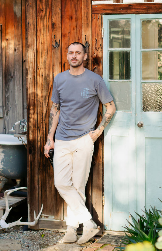 Men’s Draftsman Pant in soft washed cream showing the full-length straight-leg fit, double-front panels, and copper rivet details, styled with a Pray for Fish graphic tee.