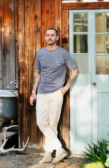 Men’s Draftsman Pant in soft washed cream showing the full-length straight-leg fit, double-front panels, and copper rivet details, styled with a Pray for Fish graphic tee.