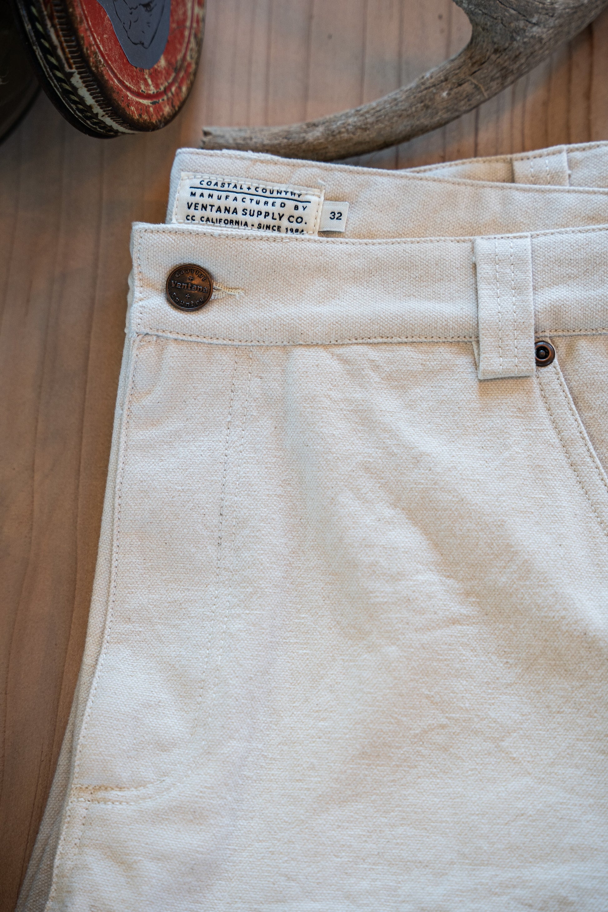 Close-up of the Draftsman Pant by Ventana Supply Co. in soft washed cream, featuring a custom-branded VNTNA metal button and authentic "Coastal + Country" woven waist label.
