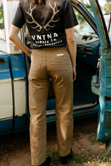 Back view of the high-waisted Lily Pant in salt washed Tobacco, featuring durable 100% cotton canvas and rear patch pockets, styled with a VNTNA Supply Co. antler graphic tee.