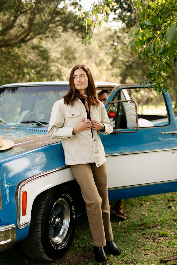 Women's high-waisted Lily Pant in salt washed Tobacco paired with the cream cotton Cove Shirt, showcasing a rugged outdoor utility look by a vintage truck.