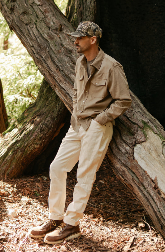 Side profile of the Tradesman Pant in softwashed cream cotton canvas, highlighting the durable triple-needle stitching and straight-leg fit, styled with a brown Western Cord shirt in a forest setting.