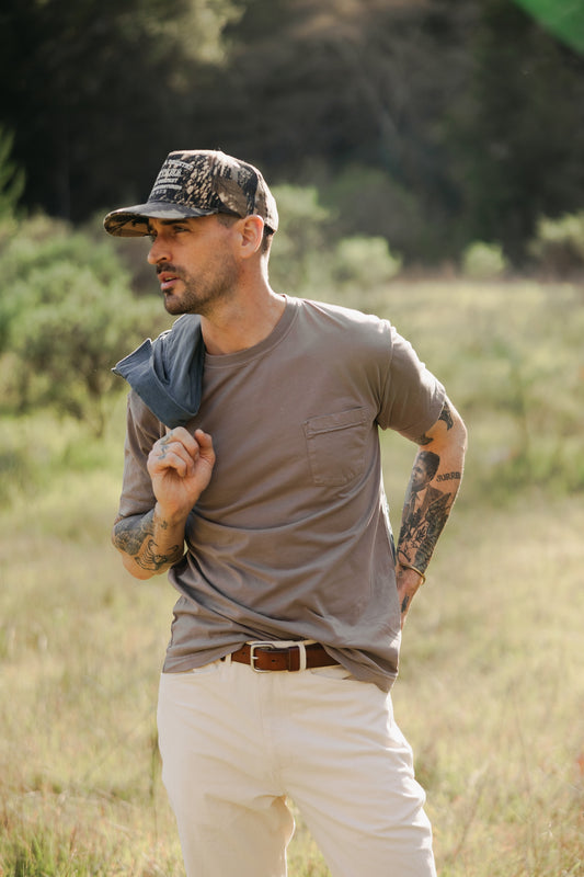 Men’s Tradesman Pant in Ivory cotton canvas, featuring a classic straight-leg fit and triple-needle stitching, styled with a pocket tee and leather belt in an outdoor field.