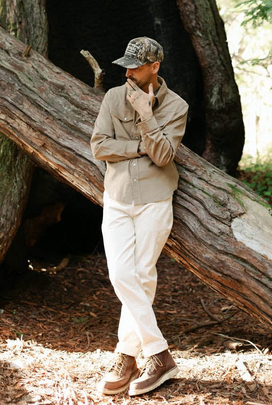 Men’s Tradesman Pant in Ivory cotton canvas, styled with the Dusty Olive Western Cord Shirt and a camo trucker hat for a rugged heritage outdoor look.