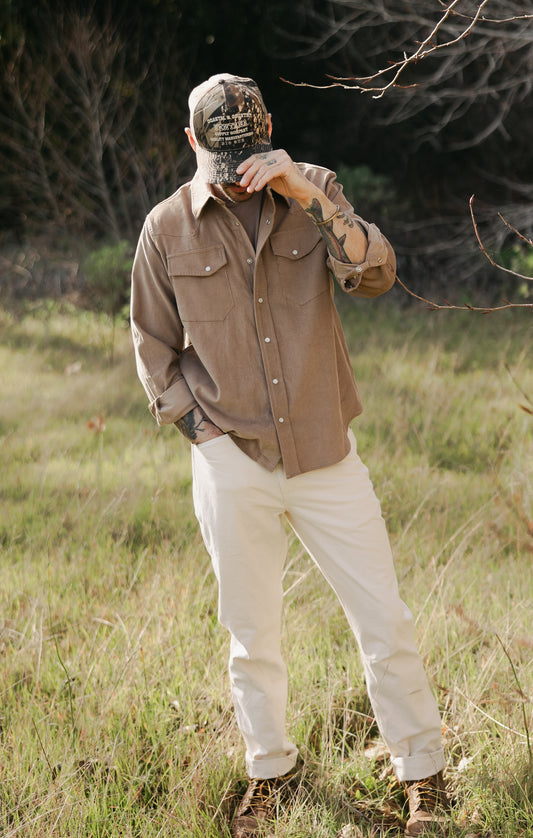 Men’s Tradesman Pant in softwashed cream cotton canvas, styled with a brown Western Cord corduroy shirt and camo trucker hat for a rugged outdoor heritage look.