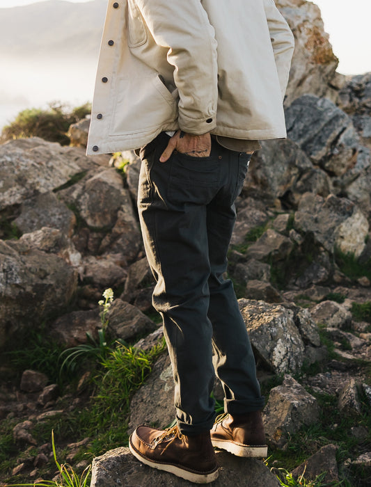 Rear view of the Tradesman Pant in salt-washed Vintage Coal, showcasing the straight-leg fit and reinforced back pockets, styled with the cream Arroyo Rancher Jacket on rocky coastal terrain.