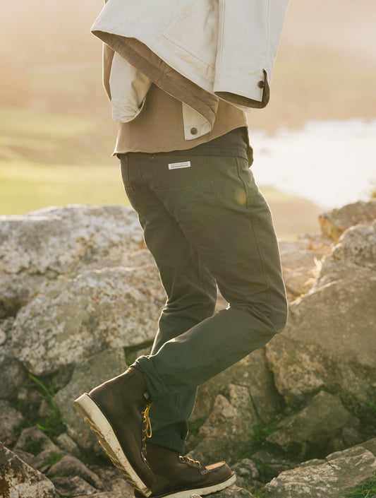 Close-up rear view of the Tradesman Pant in Vintage Coal, showcasing the Ventana Supply Co. branded woven label, durable canvas texture, and a straight-leg fit styled with work boots on a coastal trail.